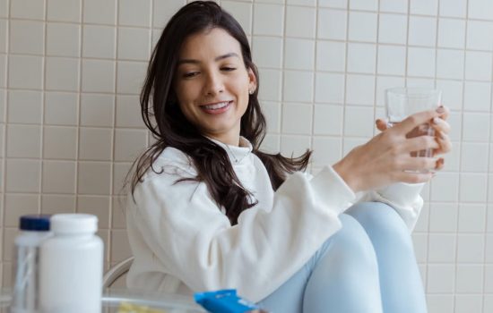 Smiling woman holding a glass while seated, with pill bottles on the table.